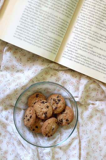 Bowl of chocolate chip cookies and open book on a bed. Flat lay.