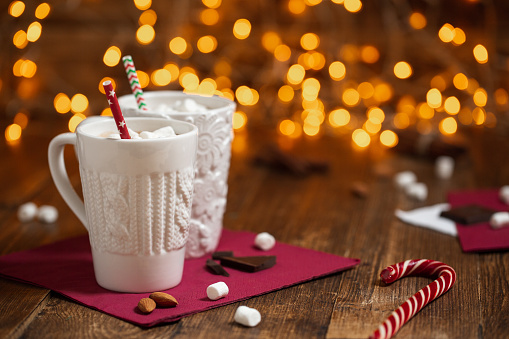 Textured white glasses on a red napkin, filled with cocoa and marshmallows, with colored paper tubes and scattered around almonds and chocolate