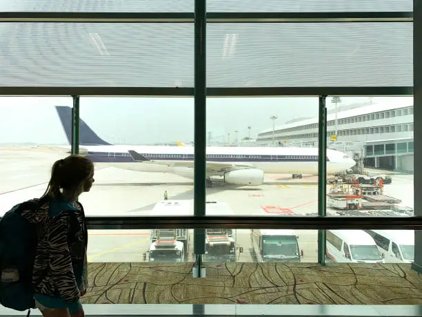 A young girl walking through an airport terminal while looking at an airplane outside on the concrete terminal.