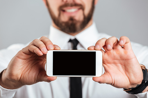 Close up of a happy businessman showing blank screen mobile phone isolated over gray background