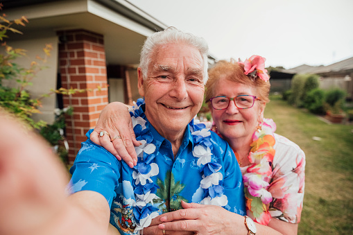 A personal perspective of a senior husband and wife standing outdoors taking a selfie together whilst wearing Hawaii themed shirts and lei's for a party in Sydney.