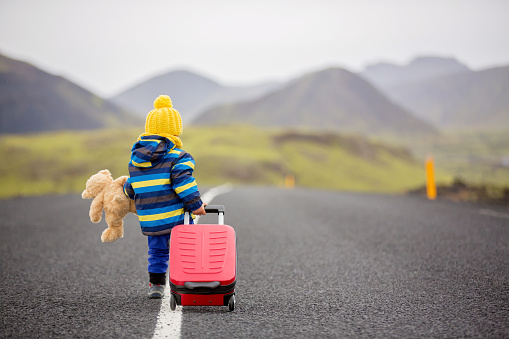 Cute toddler child, boy with teddy bear and suitcase in hand, running on a road in Iceland on a rainy autumn day