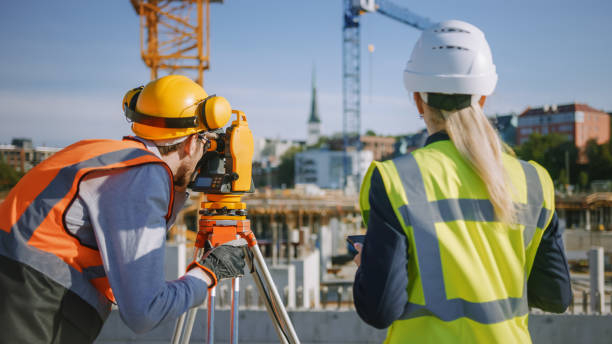 istock Construction Worker Using Theodolite Surveying Optical Instrument for Measuring Angles in Horizontal and Vertical Planes on Construction Site. Engineer and Architect Using Tablet Next to Surveyor. 1273337099