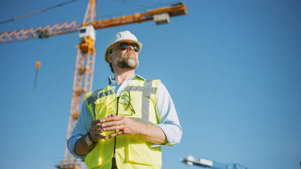 istock Confident Bearded Head Civil Engineer-Architect in Sunglasses is Standing Outside in a Construction Site on a Sunny Bright Day. Man is Wearing a Hard Hat, Shirt, Jeans and a Yellow Safety Vest. 1273337651