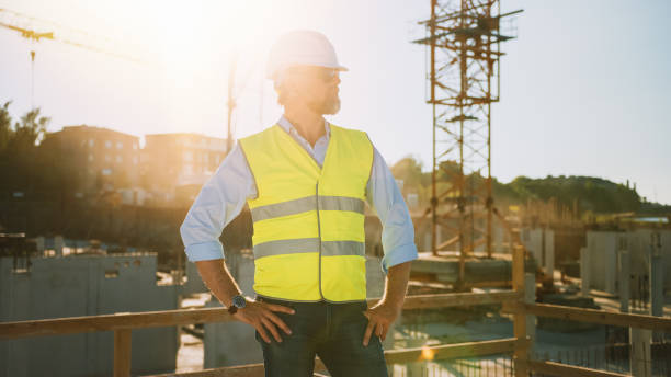 istock Confident Bearded Head Civil Engineer-Architect in Sunglasses Stands in a Construction Site on a Sunny Bright Day. Man is Wearing a Hard Hat, Shirt, Jeans and a Yellow Safety Vest. 1273337792