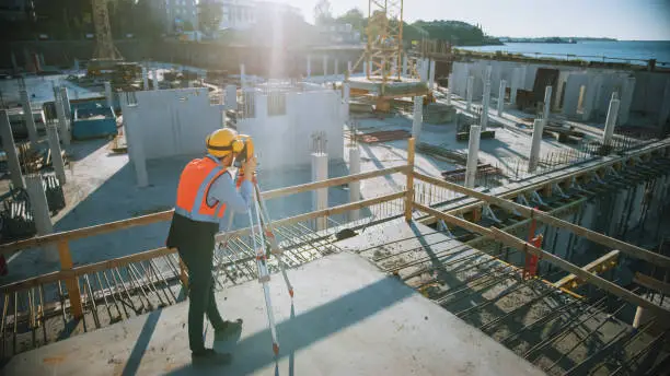 Photo of Construction Worker Using Theodolite Surveying Optical Instrument for Measuring Angles in Horizontal and Vertical Planes on Construction Site. Worker in Hard Hat Making Projections for the Building.