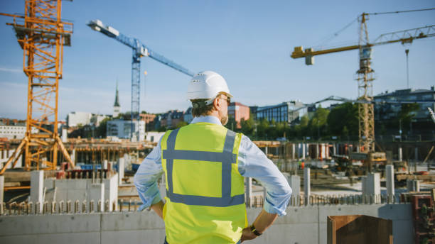 istock Confident Bearded Head Civil Engineer-Architect in Sunglasses is Standing Outside with His Back to Camera in a Construction Site on a Bright Day. Man is Wearing a Hard Hat, Shirt and a Safety Vest. 1273337821