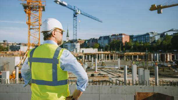 istock Confident Bearded Head Civil Engineer-Architect in Sunglasses is Standing Outside with His Back to Camera in a Construction Site on a Bright Day. Man is Wearing a Hard Hat, Shirt and a Safety Vest. 1273337875