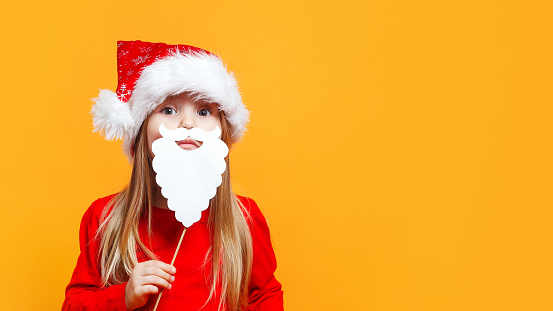 Funny child girl in Santa red hat holding paper beard on blue background.