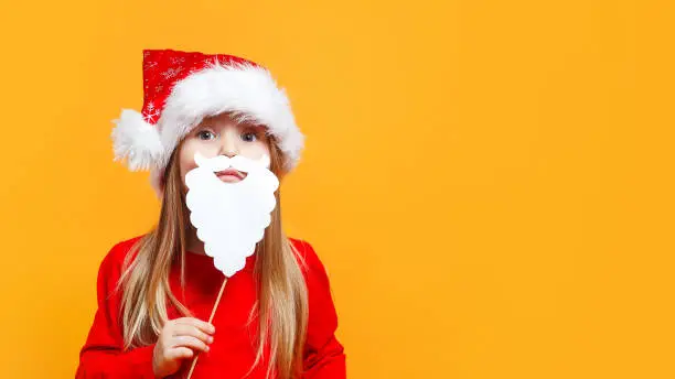 Photo of Little Santa.Funny child girl in Santa red hat holding paper beard on blue background.