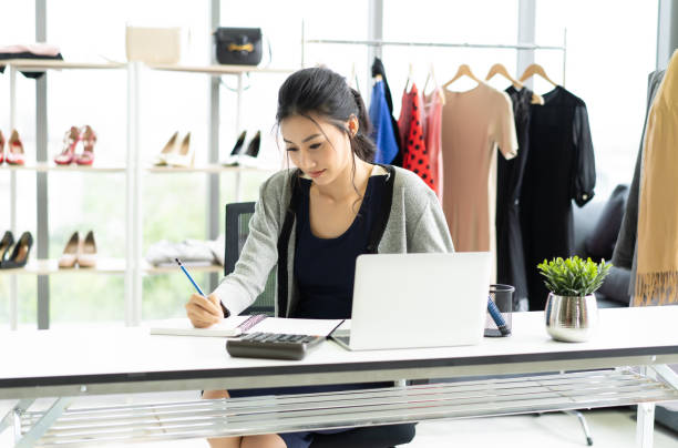 hermosa joven asiática trabajando revisando figuras y stock en su cuaderno en la tienda de ropa. startup small business owner concept. - owner boutique store retail fotografías e imágenes de stock