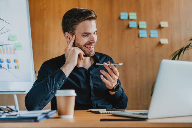 istock Caucasian young male enterpeneur talking by speakerphone and smiling while sitting on working place in modern office 1273360355