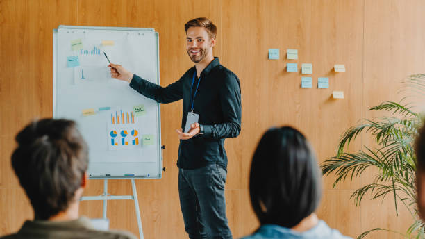 istock Caucasian businessman giving presentation to colleagues pointing on flip chart while they sitting around watching him 1273360703