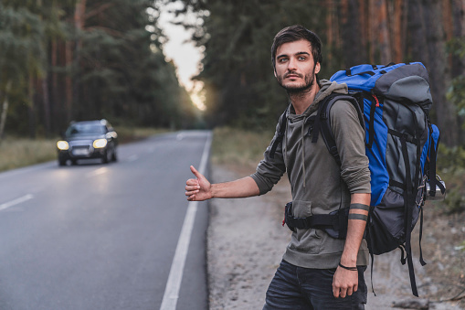 Hitchhiking concept. Young man with backpack standing on forest road while having travel trip on vacation.