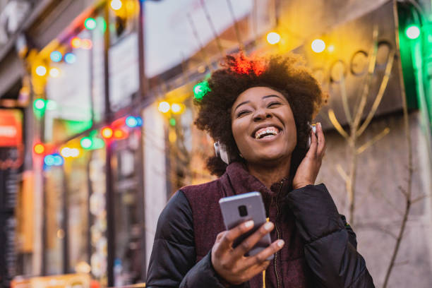 Portrait of African American woman listening music A young woman is using a mobile phone and wireless headphones outdoor during Christmas christmas excitement stock pictures, royalty-free photos & images