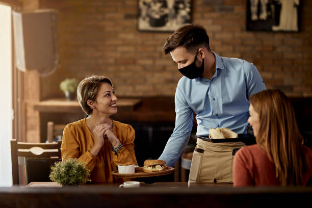 Waiter with protective face mask serving food to customers in a pub. Young waiter wearing protective face mask while serving food to his guests in a restaurant. waiter stock pictures, royalty-free photos & images