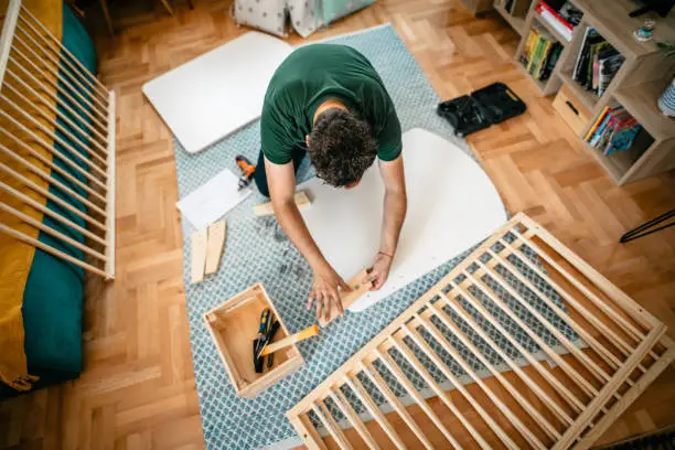 Photo of a young expectant man, future father, assembling a baby crib in the living room, having the last preparations before baby's arrival.