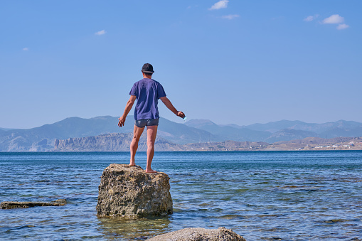 Senior asian man in shorts, t-shirt and baseball cap,holding in his hand face mask, stands alone on a stone in a sea bay over a background of mountains