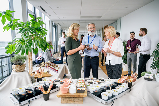 Wide angle view of Caucasian male and female business colleagues in 50s and 60s socializing at office party and enjoying gourmet dishes on buffet.