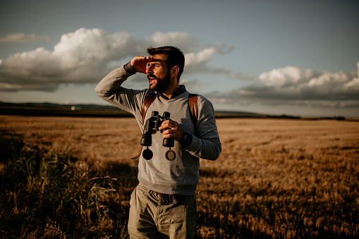 Male Traveler Looking Through Binoculars In The Distance Against The Sky.