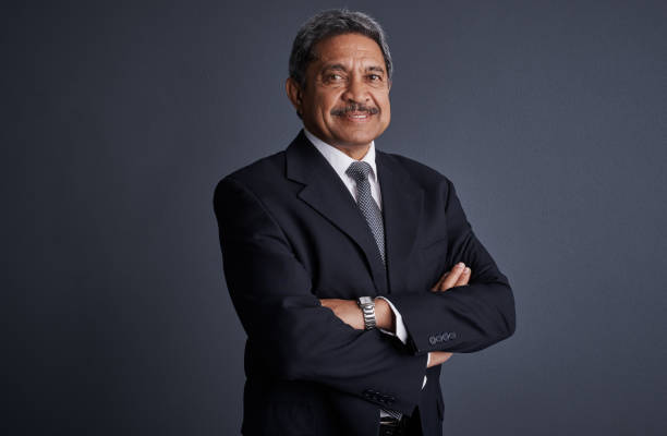 A mature Middle Eastern businessman wearing a tailored suit, tie, and white shirt, standing confidently against a bright blue studio background under soft lighting.