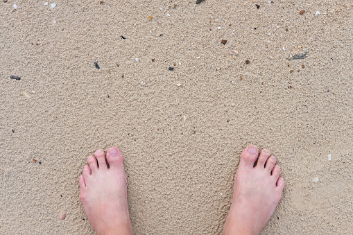 Selective focus at foot. Top view of human barefoot standing on the beach sand, small pebble and Shells with copy space.