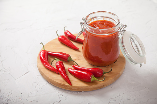 Harissa in a glass jar on a wooden stand and hot chili peppers on a white background