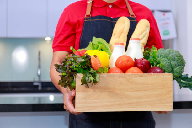istock Grocery delivery courier man in red uniform with grocery box with fresh fruit and vegetable. Deliver man in red uniform and apron handling wooden crate of food, fruit, vegetable with kitchen background. Delivery Concept 1273434586