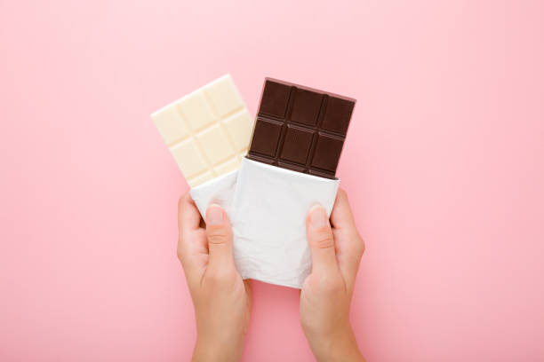 istock Young woman hands holding black and white chocolate bars on light pink table background. Pastel color. Opened packs. Sweet snack. Closeup. Point of view shot. Top down view. 1273437256