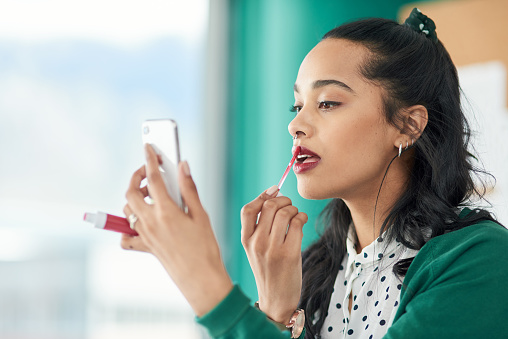 Shot of a young businesswoman applying lipstick using a smartphone in a modern office