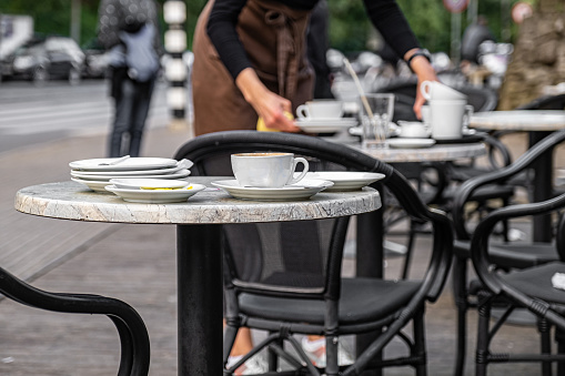 A table with uncleared dishes after visitors in a street cafe with a waitress in the background.