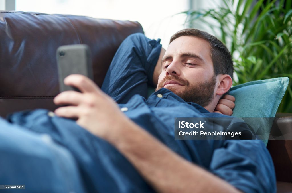 Recharge and bounce back better than ever Shot of a young man using a smartphone while relaxing on a sofa Wasting Time Stock Photo