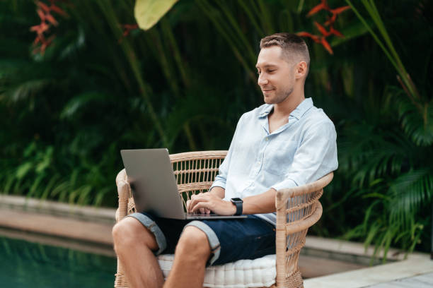 istock Young man using a laptop computer in a garden with a swimming pool. 1273442875