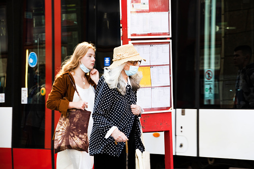 At one of the busiest tram stops in Prague 1 Center of the Capital, two females, one older wearing face mask and and a fashion straw hat and glasses, while using a walking cane. background part of tram.