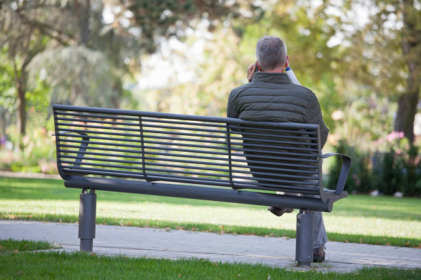 istock Mature man with face mask hanging on a ear talking on smart phone while sitting on a park bench 1273464713