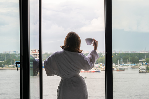 Back view of beautiful young woman in bath terry robe with cup of coffee standing and enjoying near open panoramic window of  hotel room opposite the lake.