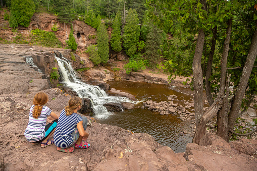 Two girls sitting at the top of a cliff near a waterfall at Gooseberry Falls State Park in northern Minnesota, USA.