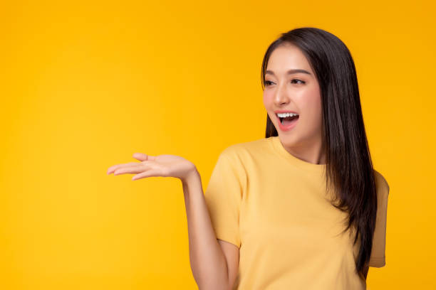 happy beautiful young asian woman with yellow background holding copy space imaginary on beauty palm for insert advertisement, happiness and smiley face. attractive beautiful girl portrait - expressive hands imagens e fotografias de stock