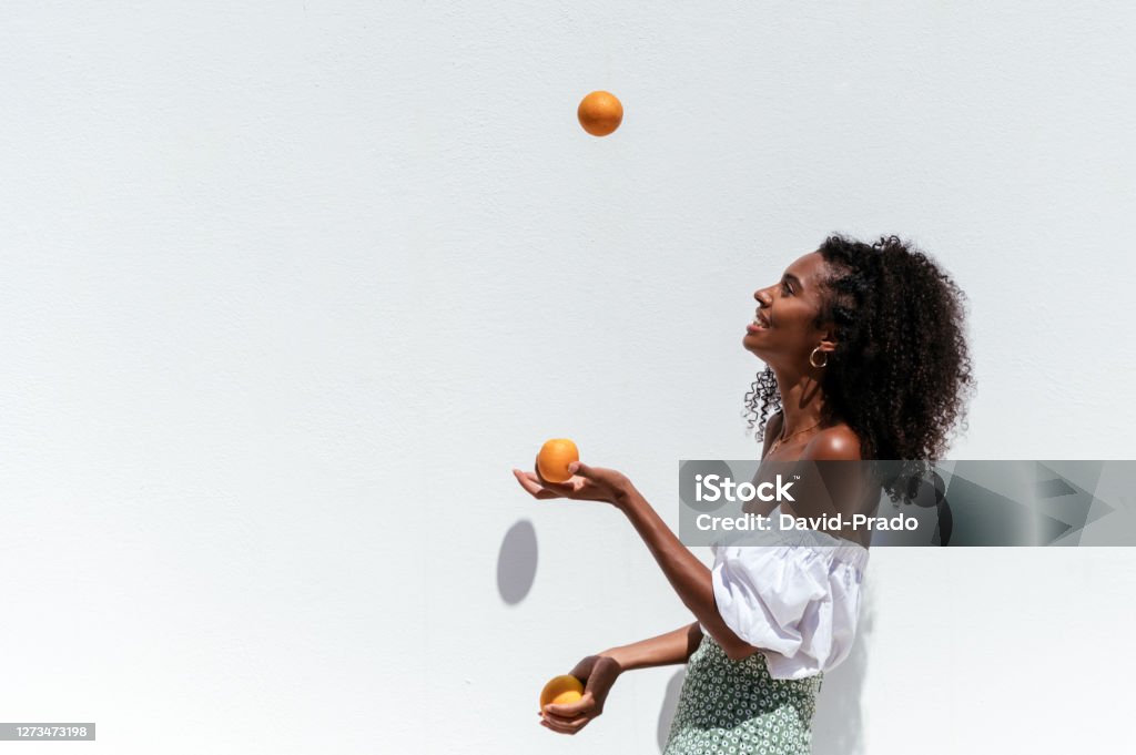 Cheerful black woman with fresh oranges Side view of happy young African American female in summer outfit juggling with ripe oranges against white wall in sunny day Juggling Stock Photo