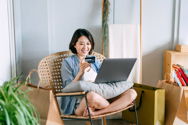 hermosa joven mujer asiática sonriente sentada en un sillón de ratán en la sala de estar en casa, comprando en línea con computadora portátil y haciendo pago móvil con tarjeta de crédito. con algunas bolsas de compras junto a ella - credit card women laptop electronic banking fotografías e imágenes de stock