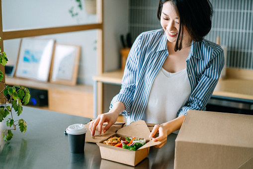 Beautiful and joyful young Asian woman opening the takeaway lunch box of fresh healthy salad, eating at home with food delivery services. Eating at home concept