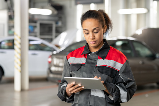 Young mixed-race female technician of contemporary car service touching display of digital tablet while reading online questions of clients