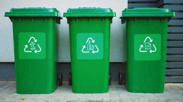 istock Green colored, plastic garbage bins, with different recycle logos on front, stacked in row against brown wood wall. Concept of waste sorting for food, paper and bottles. Saving Environment from Trash. 1273484363