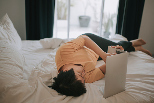 asian woman working on home lying on her bed using laptop