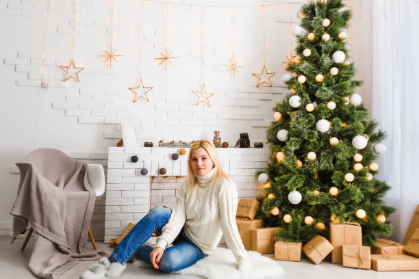 рождественская мода фото красивая девушка со светлыми волосами в уютной одежде позирует возле новогодней елки с подарками - people winter urban scene chair стоковые фото и изображения
