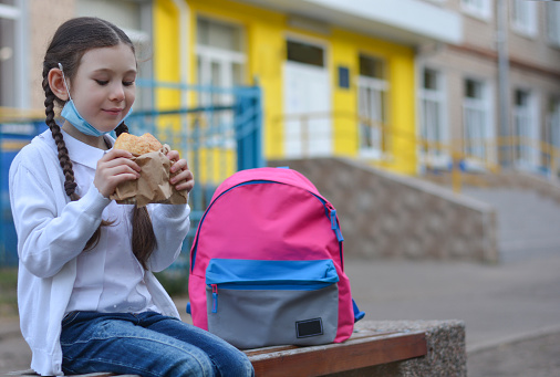 A schoolgirl with a protective medical mask, with a backpack, eating delicious pastries for lunch near the school. Back to school. New normal.