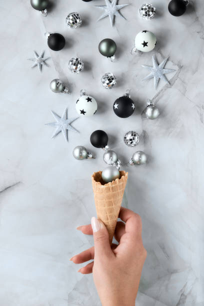 istock Creative Christmas or New Year flat lay. Hand holds ice cream waffle cone with various Xmas decorations, baubles, stars, toys in black, white, grey. Creative flat lay on marble. 1273532332
