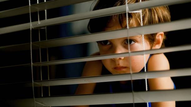 pensive little boy peeking through a window at night pensive serious little boy peeking through a window at night boy hiding behind glass stock pictures, royalty-free photos & images