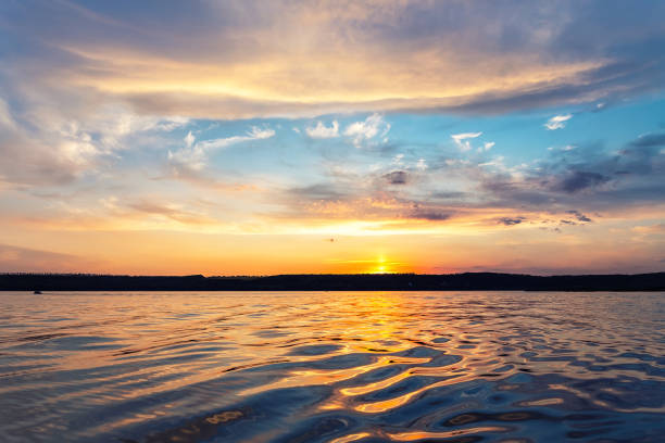 Beautiful dramatic scenic bright vibrant blue to red warm sunset evening time landscape panorama with cloudscape reflection on ripple water surface of lake or river. Horizon coast line on background. hermoso espectacular paisaje de azul brillante brillante a rojo cálido de la noche de la noche de la noche con el reflejo del paisaje nuboso en la superficie del agua ondulada del lago o el río. línea de la costa del horizonte sobre el fondo