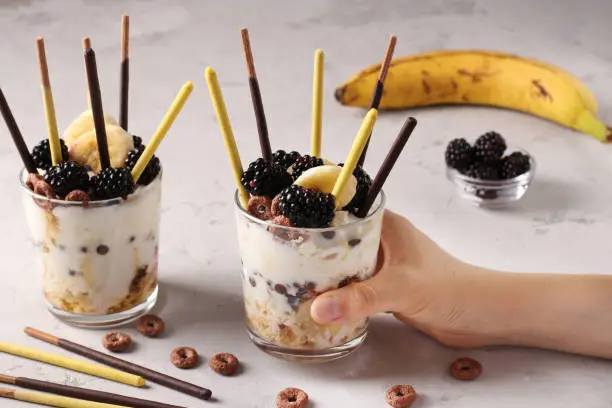 Photo of Dessert in glass with granola, chocolate, banana and blackberries decorated with sticks Pocky in female hand on gray background, Closeup